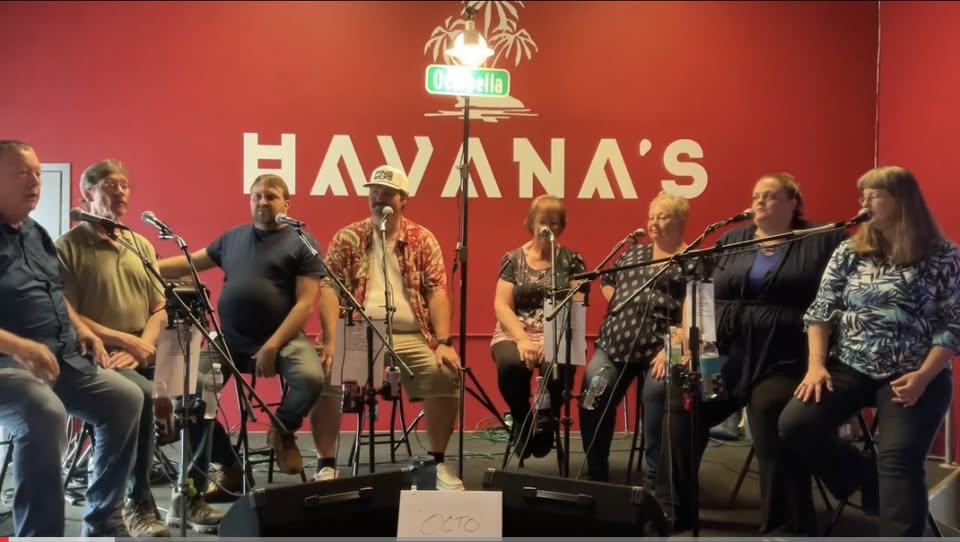 Eight people sit onstage with microphones at HAVANA'S, a red-walled venue with a palm tree sign above them. They appear to be preparing to perform or sing together.
