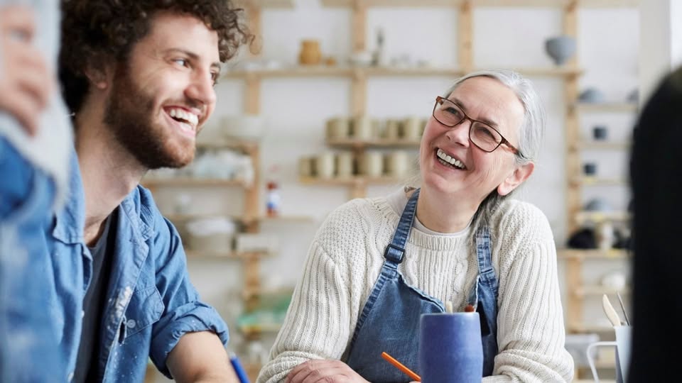 Two people, one older woman wearing glasses and an apron, and a younger man with curly hair and a beard, smile and talk together in a bright pottery studio with shelves of ceramics in the background.