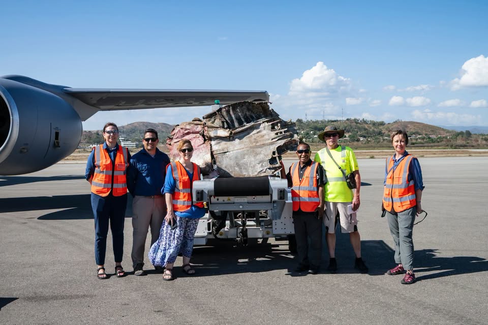 Six people in safety vests stand on an airport tarmac in front of an airplane wing and a large piece of twisted debris on a cart, with hills and a blue sky in the background.