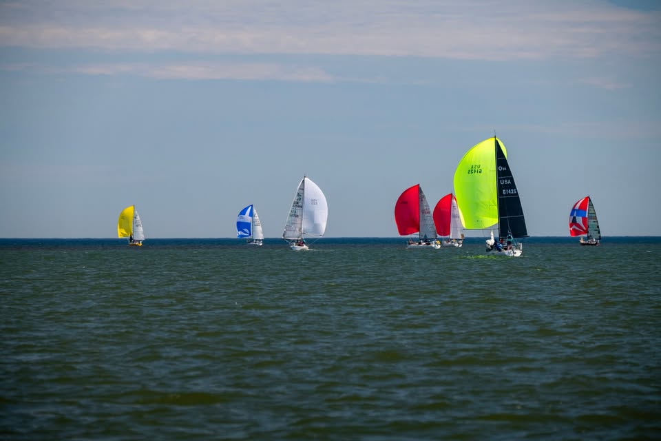 Several sailboats with colorful sails, including yellow, blue, white, red, and neon green, race across a calm body of water under a partly cloudy sky.