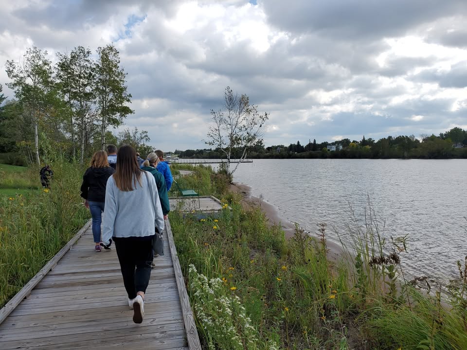 A group of people walk along a wooden boardwalk beside a lake, surrounded by tall grass and trees under a cloudy sky.