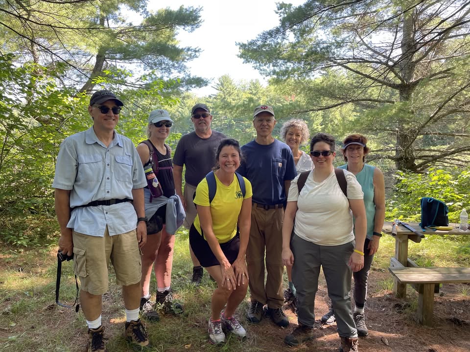 A group of eight adults stands and smiles on a forest trail, surrounded by trees and greenery. Some wear hats, sunglasses, or backpacks, with a wooden bench partly visible on the right.