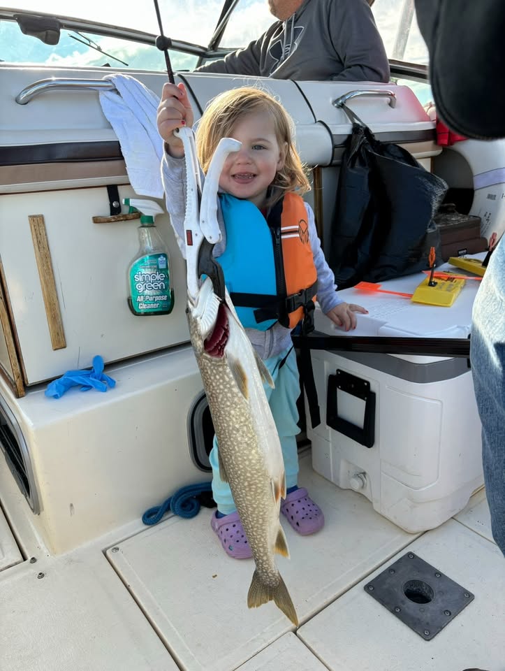 A young child in a life jacket proudly holds up a large fish on a boat, smiling widely. Boating supplies and cleaning spray are visible behind her.