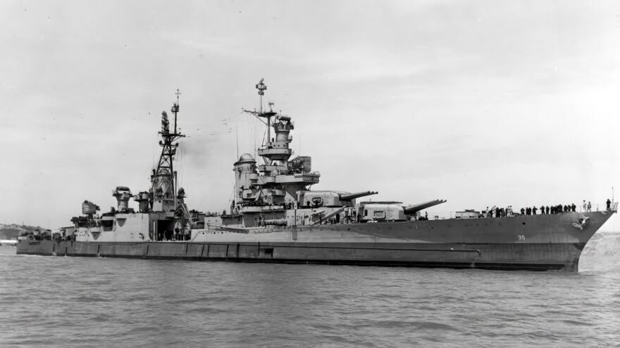 A large World War II battleship with multiple gun turrets sits in calm water, viewed from the side, with crew members visible along the deck under a cloudy sky.