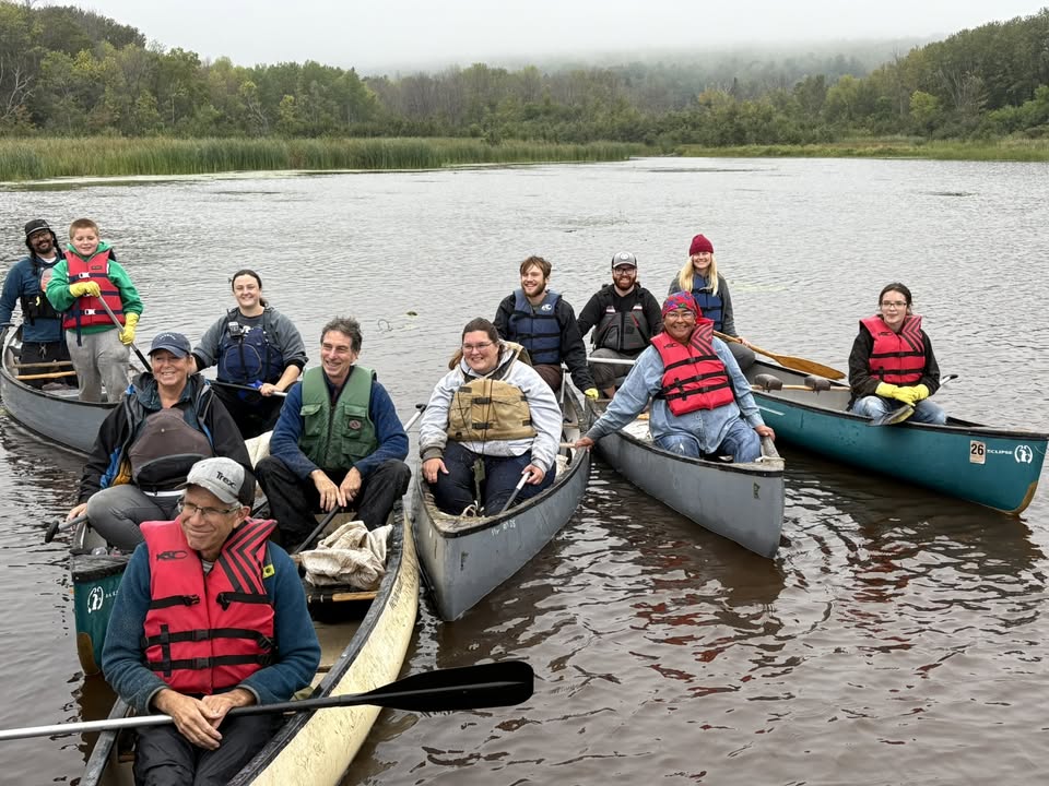A group of people wearing life jackets sit in three canoes on a calm lake, smiling at the camera. Green trees and reeds are visible in the background under an overcast sky.
