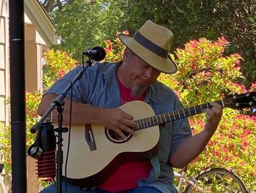 A person wearing a hat and plaid shirt plays an acoustic guitar outdoors, seated near a microphone, with green and red foliage in the background.