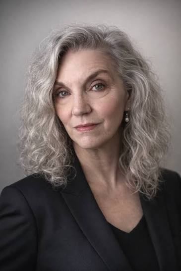 A confident older woman with curly gray hair, wearing a black blazer and pearl earring, looks directly at the camera with a neutral expression against a plain background.