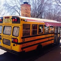 A yellow school bus converted into a food truck with a red and white striped awning, parked by the curb under leafless trees.