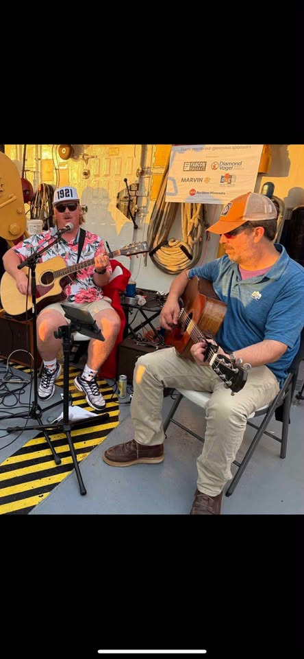 Two men sit on chairs playing acoustic guitars. One wears a floral shirt and sunglasses, the other wears a blue shirt and cap. They perform together outdoors near a sign and equipment.