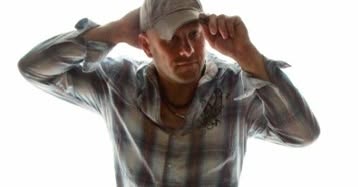 A person wearing a plaid shirt and a cap tilts the brim of the cap forward with one hand, while the other hand rests on the back of their head, posing against a bright white background.