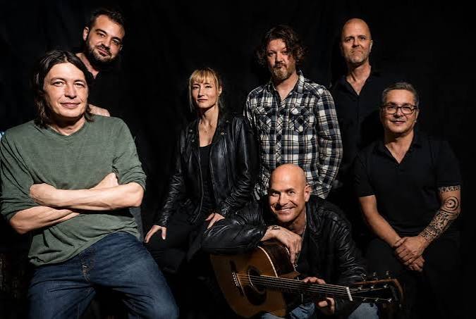 Seven adults pose together against a dark background; one holds an acoustic guitar. Most are smiling or neutral. They wear casual clothing, with one person wearing a plaid shirt and another wearing a black leather jacket.