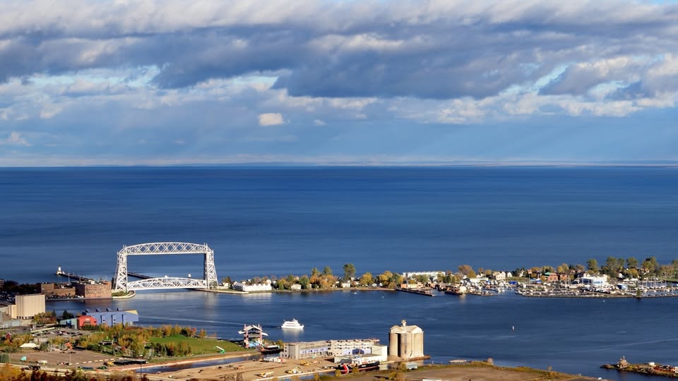A view of a large blue lake with a white lift bridge, boats, and buildings along the shoreline under a partly cloudy sky.