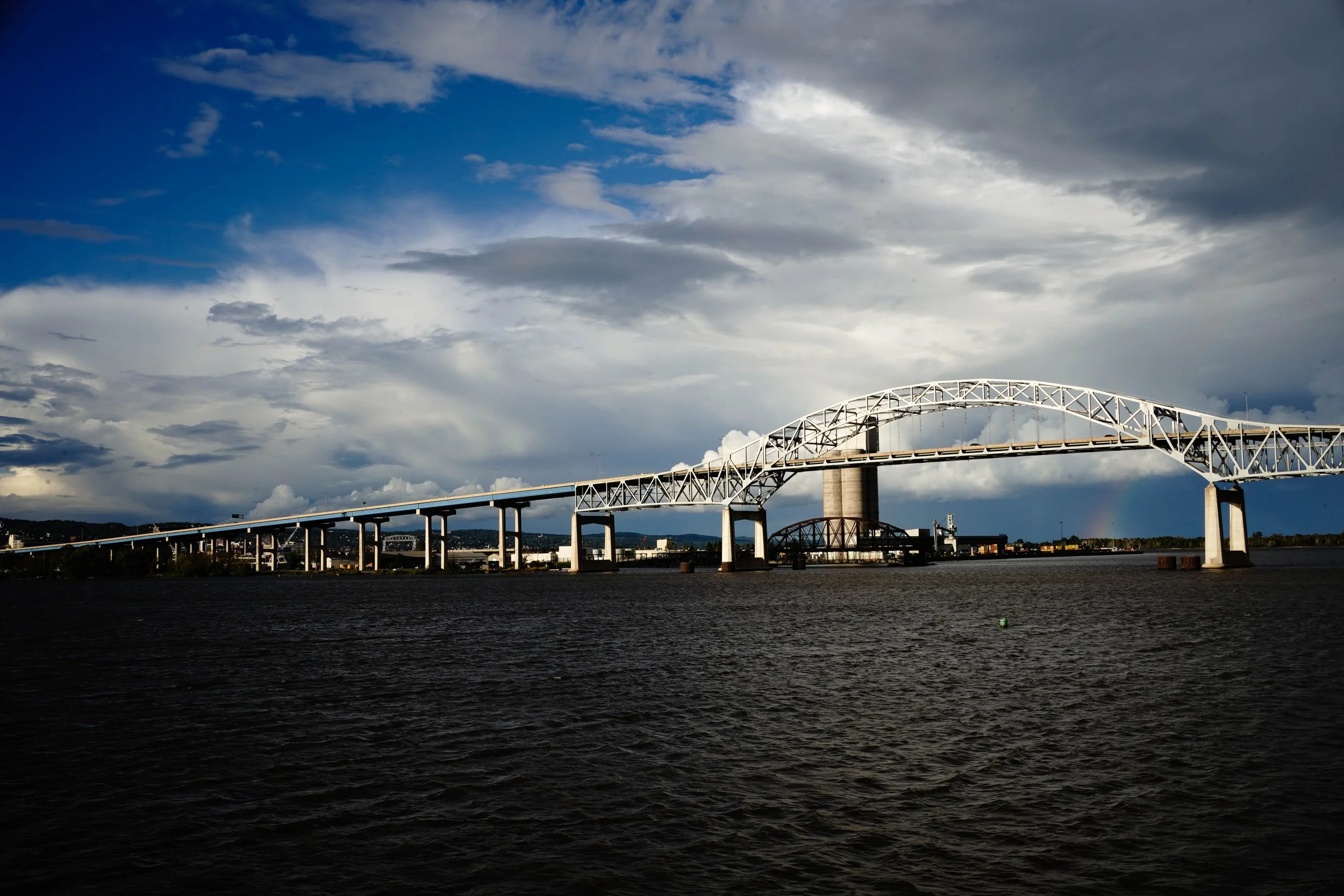 A large white arch bridge spans across a wide river under a dramatic, partly cloudy sky with industrial buildings visible in the background.