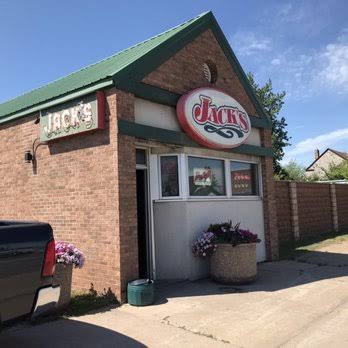 A small brick building with green roof trim has two signs reading “Jack’s.” There are potted flowers by the entrance, a pickup truck parked nearby, and a partly cloudy sky overhead.