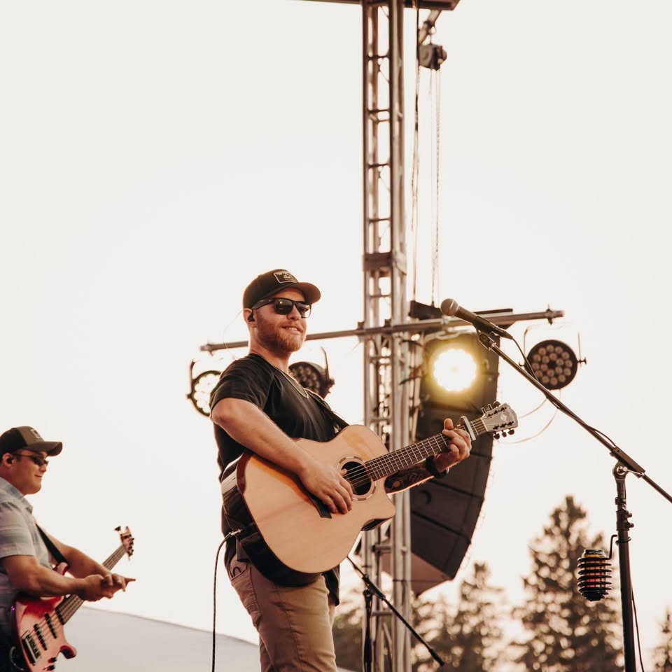 A musician wearing sunglasses and a hat plays an acoustic guitar on stage outdoors. Another musician with a bass guitar is visible in the background. Stage lights and trees are behind them.