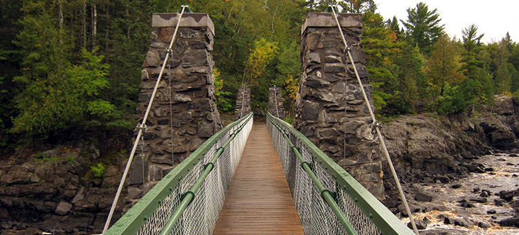 A narrow wooden suspension bridge with stone towers crosses a rocky river, surrounded by dense green trees and forest.