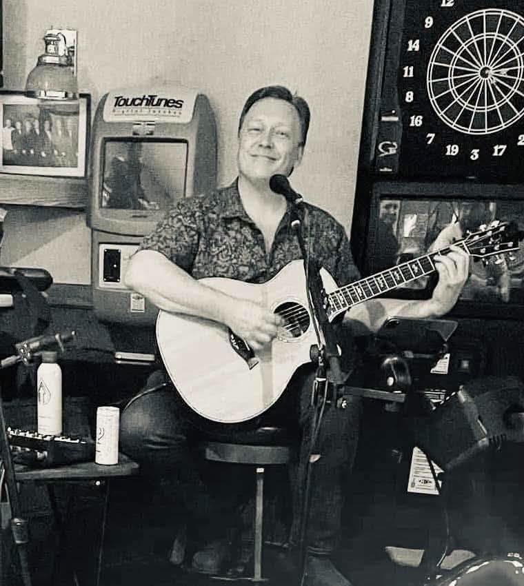 A man sits and smiles while playing an acoustic guitar. Behind him are a dartboard, a TouchTunes jukebox, and framed photos. Two drink cans are visible on a small table nearby.