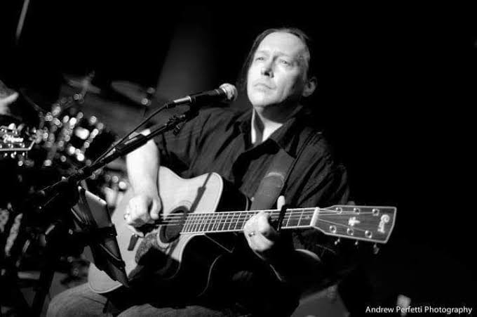 A man sits on stage playing an acoustic guitar and singing into a microphone. The photo is in black and white, with a soft focus on the background, highlighting the musician’s focused expression.