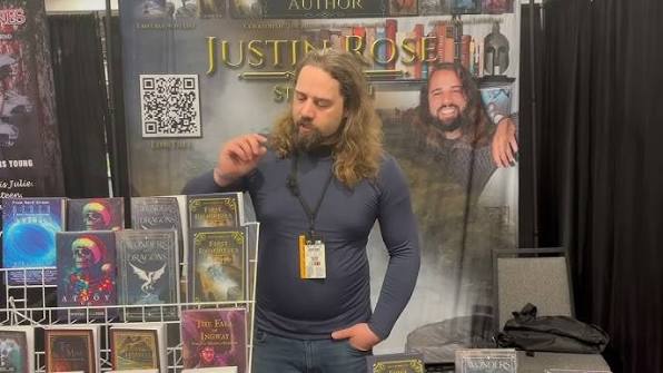A man with long hair and a beard stands at a book booth, surrounded by fantasy novels. A large banner with his photo and name is behind him. He appears to be thinking or looking down while holding something in his hand.