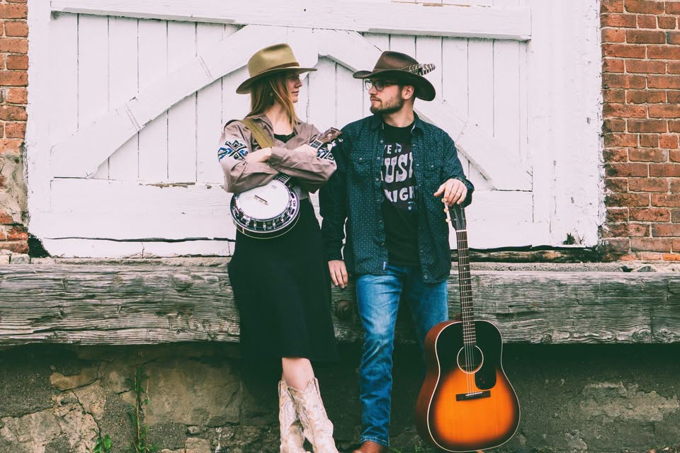Two people in cowboy hats stand by a rustic wooden door. One holds a banjo, wearing boots and a patterned shirt, while the other, in jeans and glasses, stands with an acoustic guitar resting on the ground.