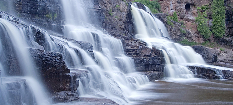 A multi-tiered waterfall cascades over dark rocks with lush green plants growing on the cliffside, and water flows into a calm pool below.