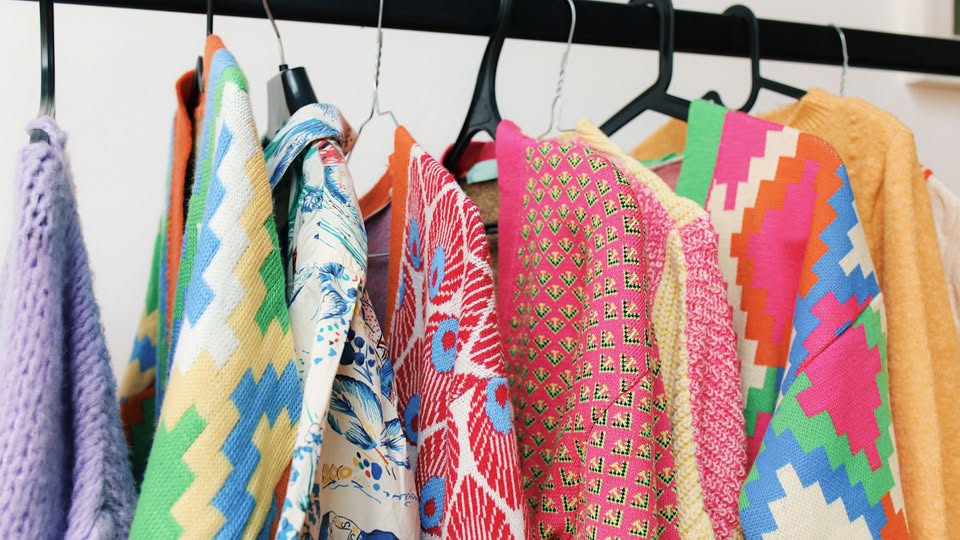 Colorful patterned sweaters and shirts on black hangers hang from a clothing rack. The garments feature bold geometric, floral, and abstract designs in bright colors like yellow, red, blue, pink, and green.