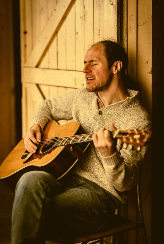 A man sits on a chair playing an acoustic guitar and singing with his eyes closed, wearing a beige sweater and jeans, next to a wooden door.