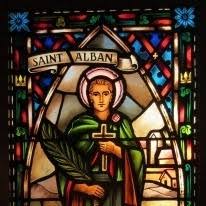 Stained glass window depicting Saint Alban holding a cross and a palm leaf, with a banner above reading “Saint Alban,” set against a colorful background with floral designs.