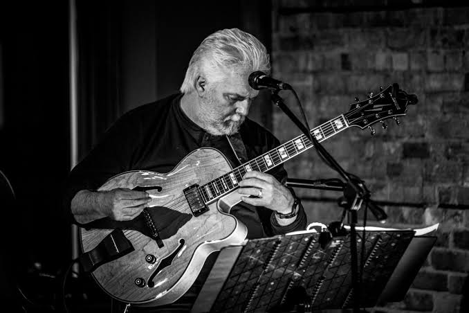 A grayscale image of an older man with white hair playing an electric guitar on stage, seated behind a microphone, with sheet music in the foreground and a brick wall in the background.