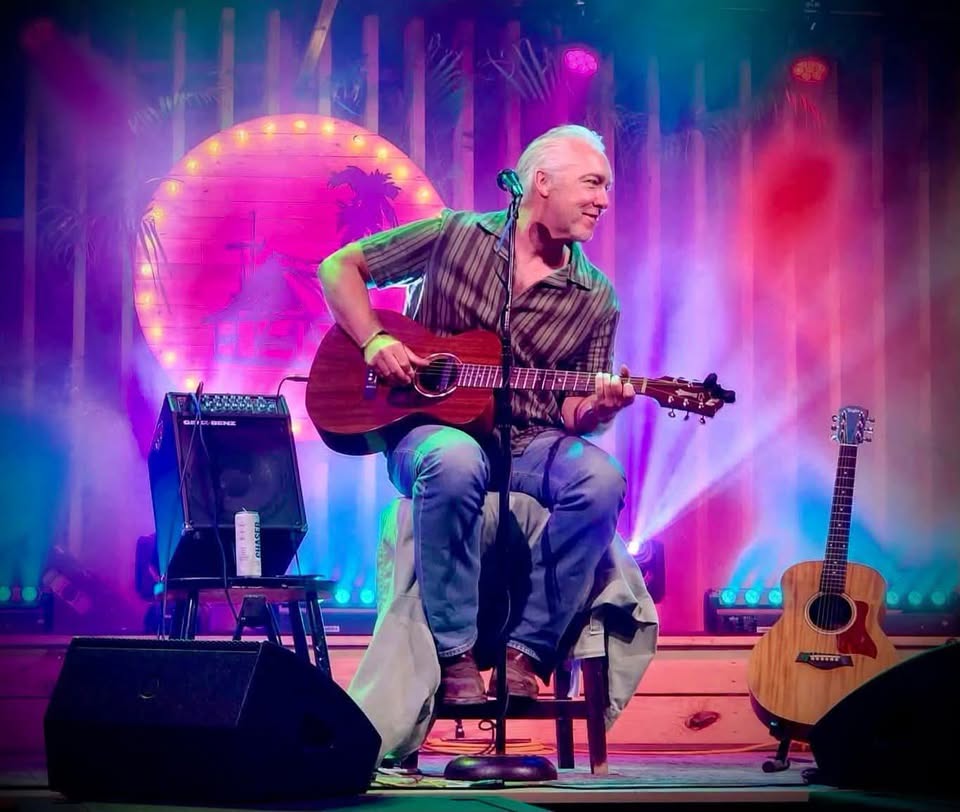A man with gray hair sits on a stool playing an acoustic guitar on stage, smiling. Colorful lights and a neon palm tree backdrop illuminate the scene. An amplifier and another guitar are beside him.