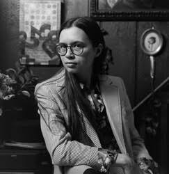 Black and white photo of a person with long hair and glasses wearing a striped blazer, sitting in a room with framed art and flowers in the background.