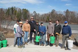 A group of nine people stands outdoors near a river, holding green buckets and litter pickers, with trees and a cloudy sky in the background.
