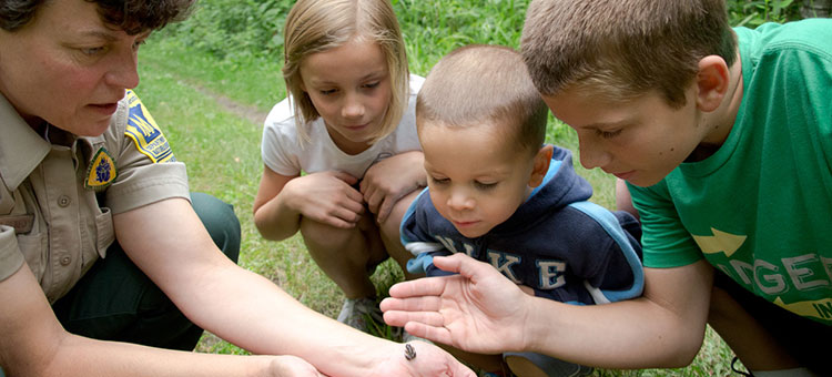 A park ranger shows something small in her hand to three curious children outdoors on a grassy path.