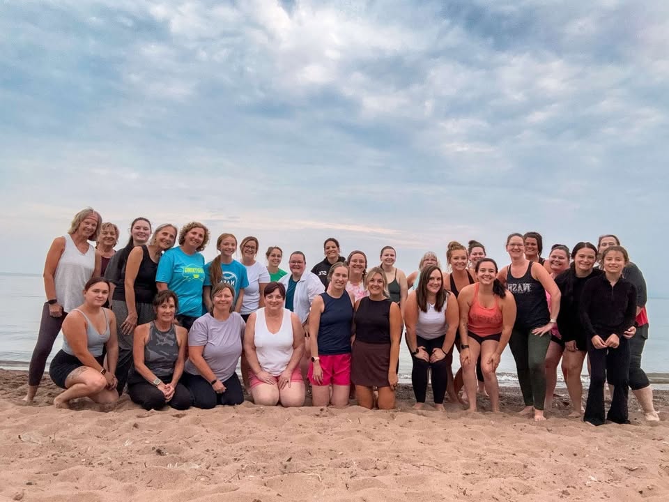 A large group of women pose together on a sandy beach near calm water under a cloudy sky, most smiling at the camera.