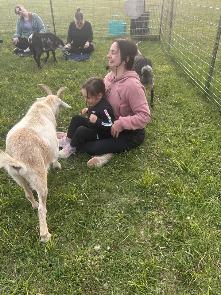 A woman in a pink hoodie sits on the grass holding a smiling child as a goat approaches them. Other people and goats are nearby in a fenced grassy area.