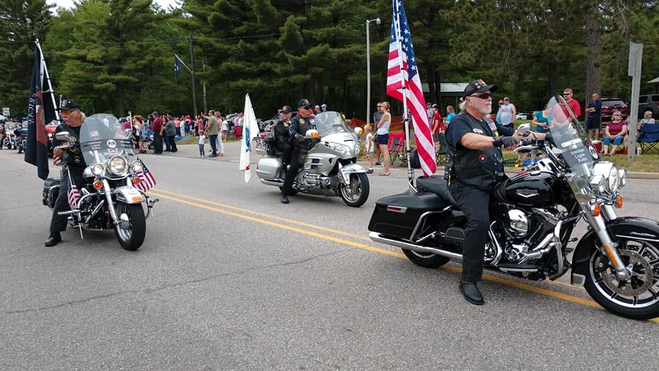 Motorcycle riders carry American flags in a parade, passing crowds lined along a tree-lined street. The riders wear black vests and helmets, and several motorcycles have small flags attached.