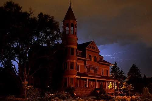 A large, dark Victorian house stands under a stormy night sky, illuminated by flashes of lightning behind it, with trees silhouetted in the foreground.