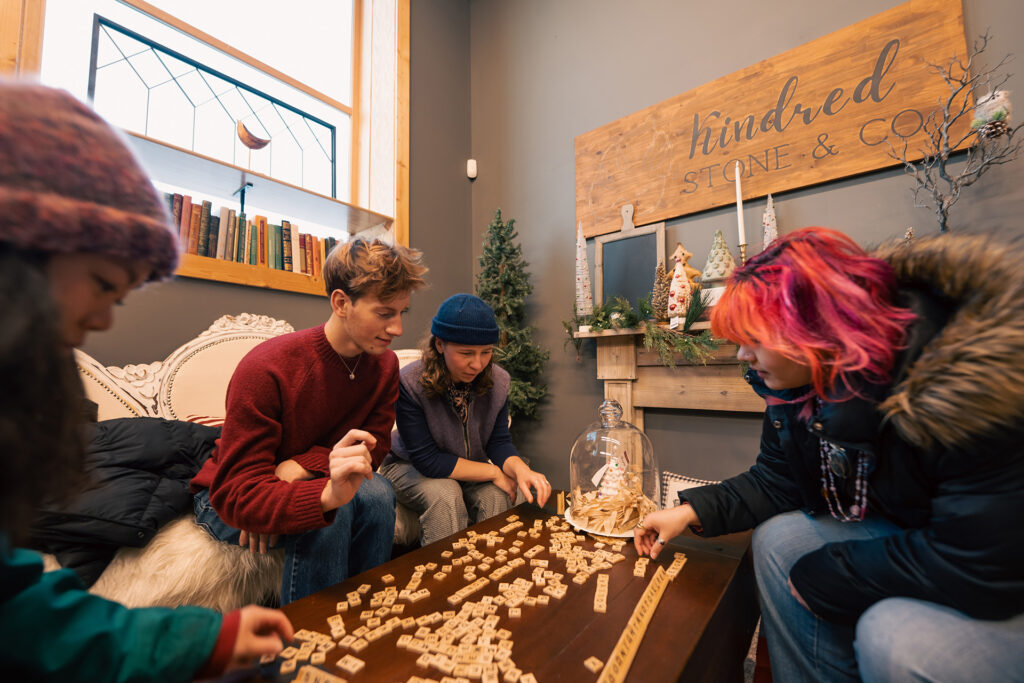 Four people sit around a coffee table playing a word tile game in a cozy room adorned with winter hats, holiday decorations, and a wooden sign reading “kindred STONE & CO.” on the wall behind them—a perfect scene to inspire your holiday shopping in Superior, Wisconsin.