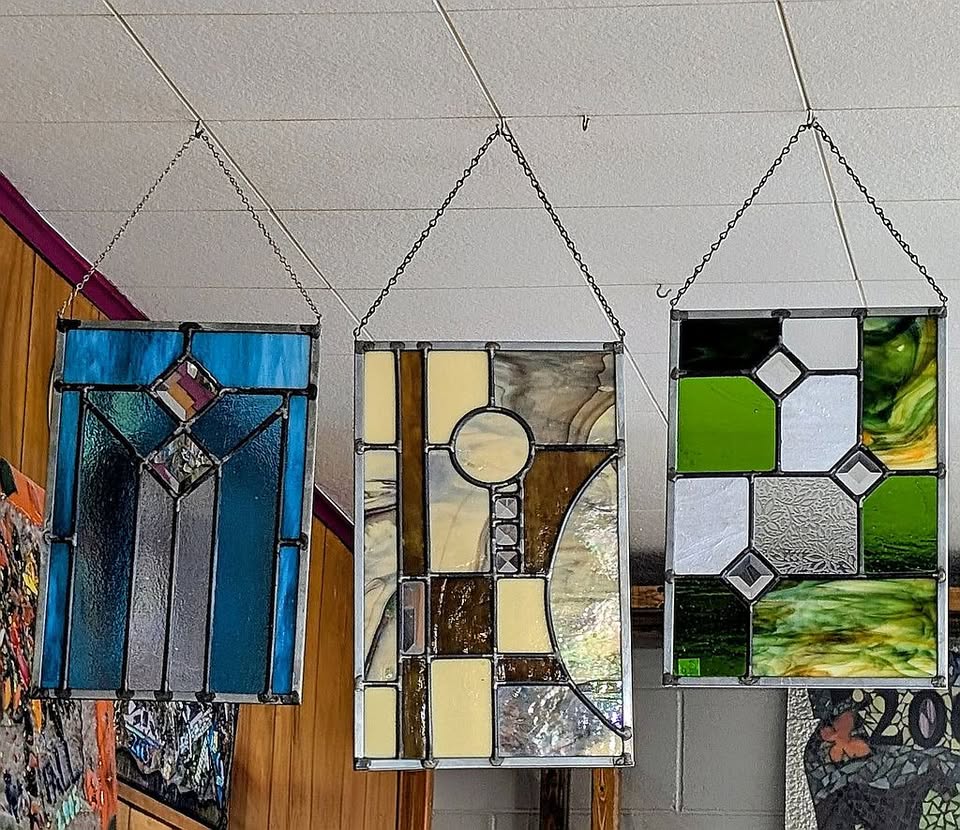 Three stained glass panels hang from chains. The panels feature geometric designs in blue, cream, gray, green, and clear glass, displayed indoors against a wood-paneled and white-tiled ceiling background.