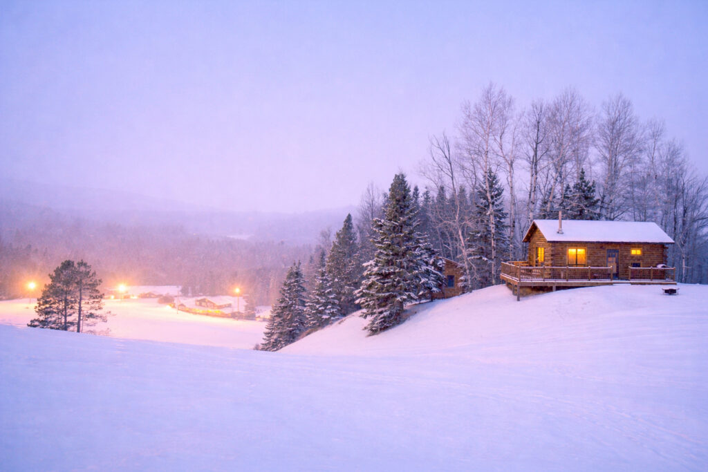 A small wooden cabin with glowing windows sits on a snowy hill surrounded by trees at dusk, evoking the cozy charm of holiday shopping in Superior, Wisconsin. Distant lights from other buildings glow in the snowy landscape under a soft, light purple sky.