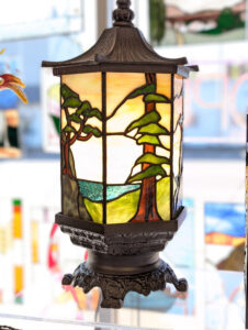 Stained glass lamp with a nature scene, featuring trees and a lake, sits on a reflective surface. The lamp has a dark ornate metal base and frame, with bright daylight in the background—a perfect accent piece to discover during holiday shopping in Superior Wisconsin.