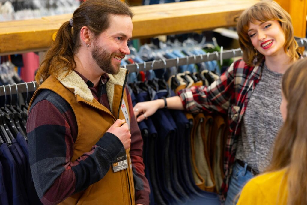 Three people are talking and smiling in a clothing store during holiday shopping in Superior, Wisconsin. A man with long hair and a beard stands near a clothing rack, while two women, one in plaid, laugh and chat with him. Shirts hang neatly on hangers behind them.