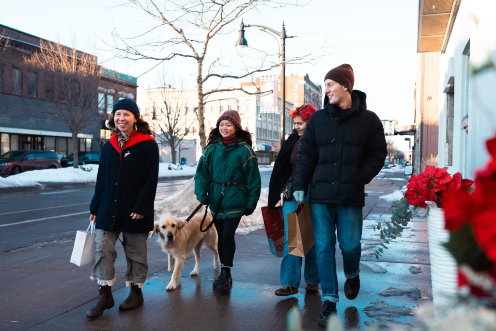 Four people and a dog walk on a snowy city sidewalk in Superior, Wisconsin, carrying shopping bags and dressed in winter clothes, capturing the spirit of holiday shopping. A vase of red flowers decorates the right side of the image.