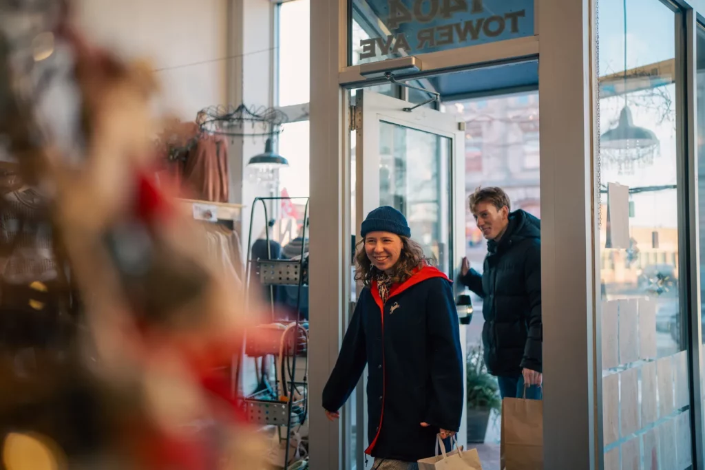 A smiling woman in a blue hat and red scarf enters a store holding shopping bags, followed by a man. The scene captures the spirit of holiday shopping in Superior, Wisconsin, with bright and festive atmosphere, and blurred holiday decorations sparkling in the foreground.