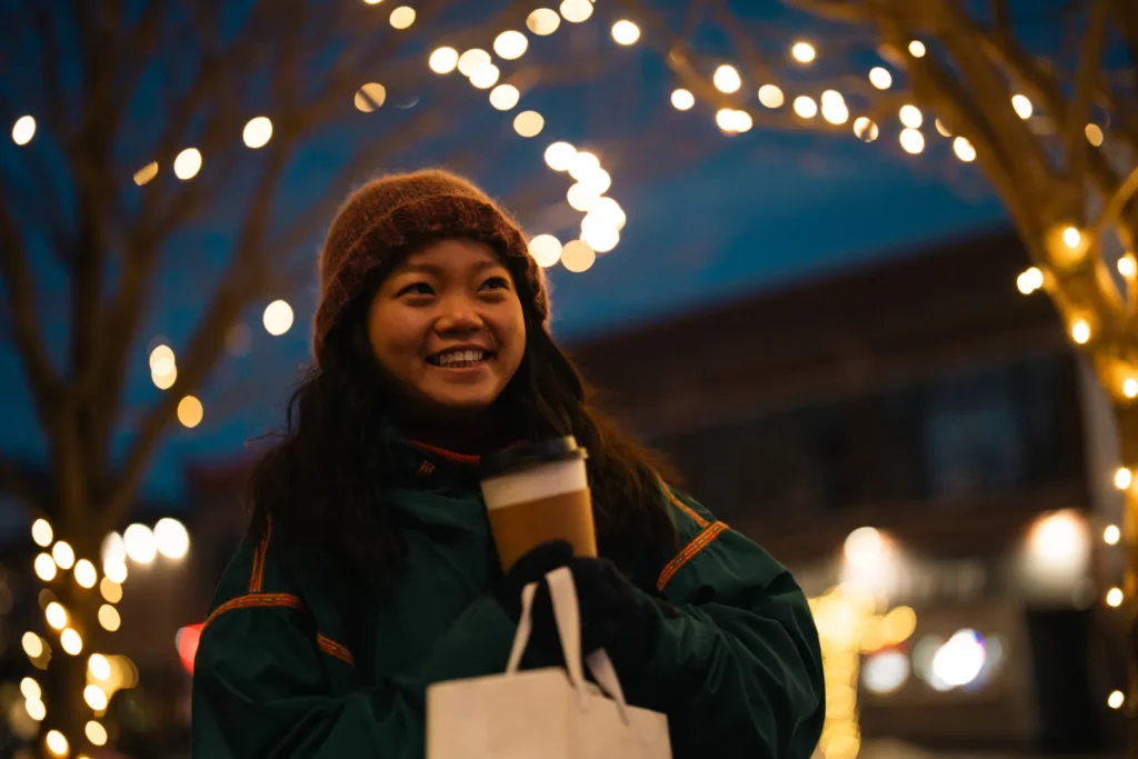 A woman in a winter coat and knit hat smiles, holding a coffee cup and shopping bag, standing outside at night with festive string lights in the background—capturing the joyful spirit of holiday shopping in Superior, Wisconsin.