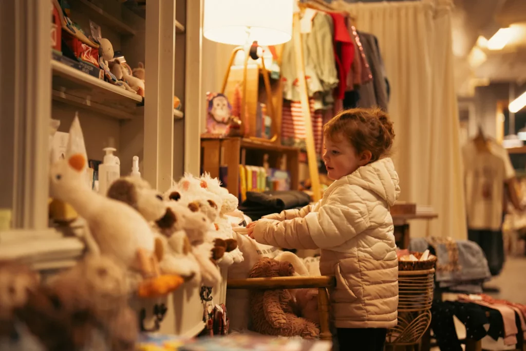 A young child in a cream jacket smiles and reaches for stuffed animals on a shelf in a warmly lit store, surrounded by toys and clothes—capturing the joyful spirit of holiday shopping in Superior, Wisconsin.