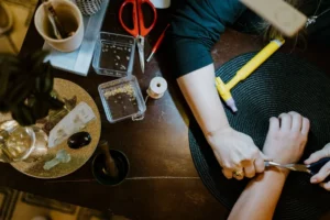 Person crafting jewelry at a table, using pliers on a bracelet—perhaps creating the perfect unique gift for holiday shopping in Superior, Wisconsin. Scissors, beads, a yellow tool, and various materials are spread around on a dark tabletop.