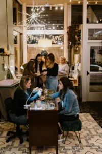 Two women sit at a small table in a cozy, warmly lit shop during holiday shopping in Superior, Wisconsin, while several people gather and talk in the background near large windows decorated with string lights.