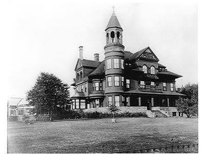 Black and white photo of a large Victorian-style house with a prominent round tower, peaked rooftops, and multiple chimneys, set on a spacious lawn with a tree in the foreground.