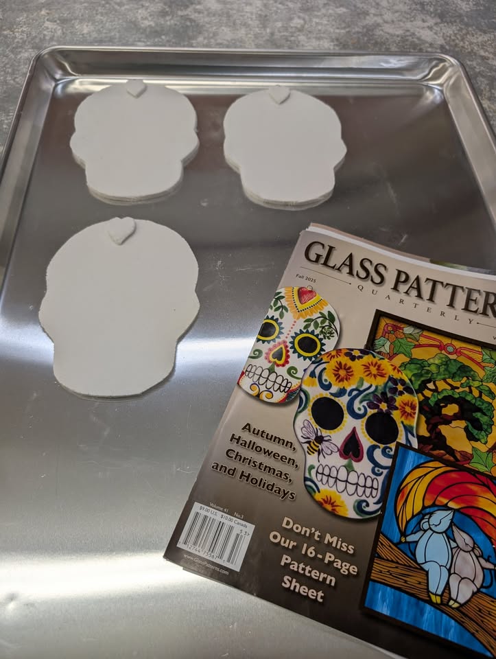 Three blank skull-shaped objects on a metal tray next to a magazine featuring colorful sugar skull artwork and stained glass designs.